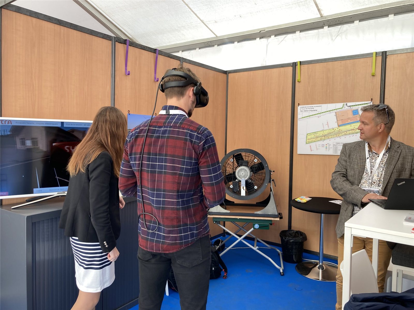 Un apprenant en casque VR face au ventilateur, la vue retransmise sur l'écran du stand